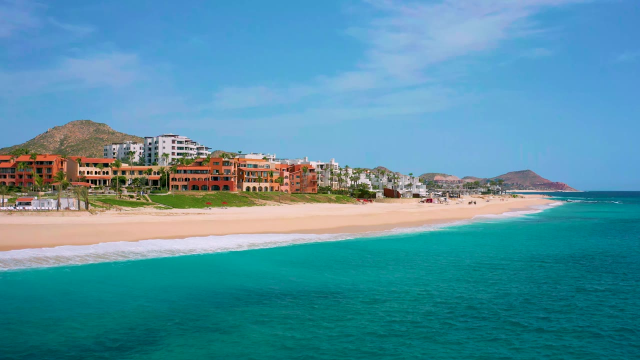 Aerial coastline in Desert Park Natural Reserve, Baja California Sur, showing blue-green water, a pale beach, red-orange resort buildings, white hotel blocks, hills, and no visible people.