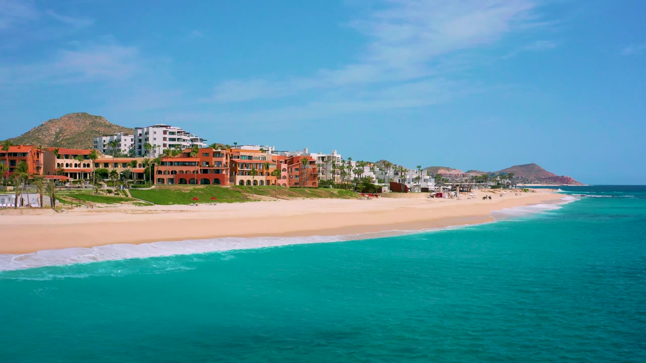 Aerial frame from Desert Park Natural Reserve, Baja California Sur, with small surf washing onto sand below resort buildings, hillside terrain behind the shore, and no visible people.