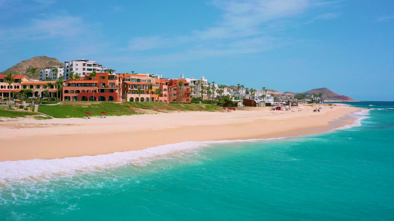 Aerial view of Desert Park Natural Reserve coastline in Baja California Sur, with turquoise surf in the foreground, terracotta resort buildings above the sand, distant hills, and no visible people.