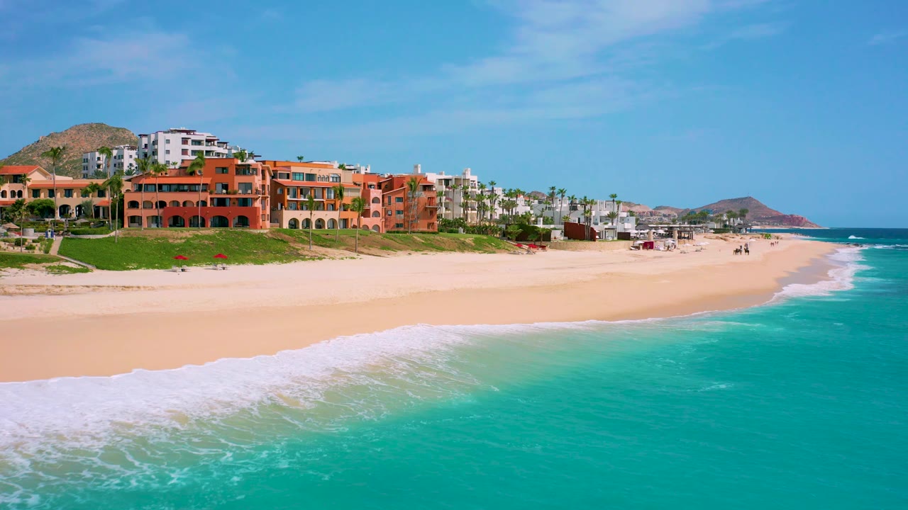 Aerial view over the Baja California Sur resort beachfront, with foam reaching farther across the pale sand, terracotta buildings along the dune, low hills behind them, and no visible people.