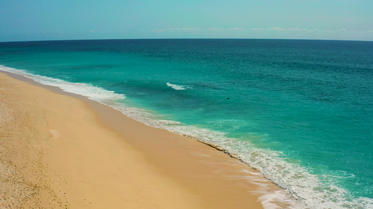 Opening aerial frame of Desert Park Natural Reserve coastline in Baja California Sur, with golden sand at left, turquoise surf at right, a dark blue horizon, and no visible people.