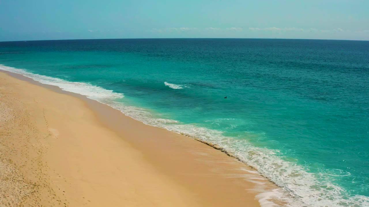 Aerial view one second into the clip shows the Baja California Sur beach running diagonally beneath clear blue water, with small foam breaks and no visible people.