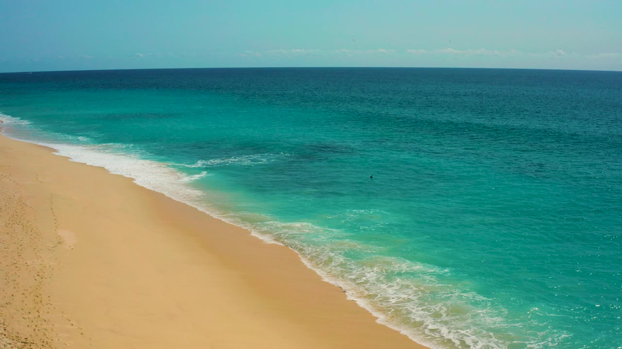 Aerial view in Baja California Sur, Mexico, with golden sand at left, turquoise water at right, white foam along the diagonal shoreline, and no visible people.
