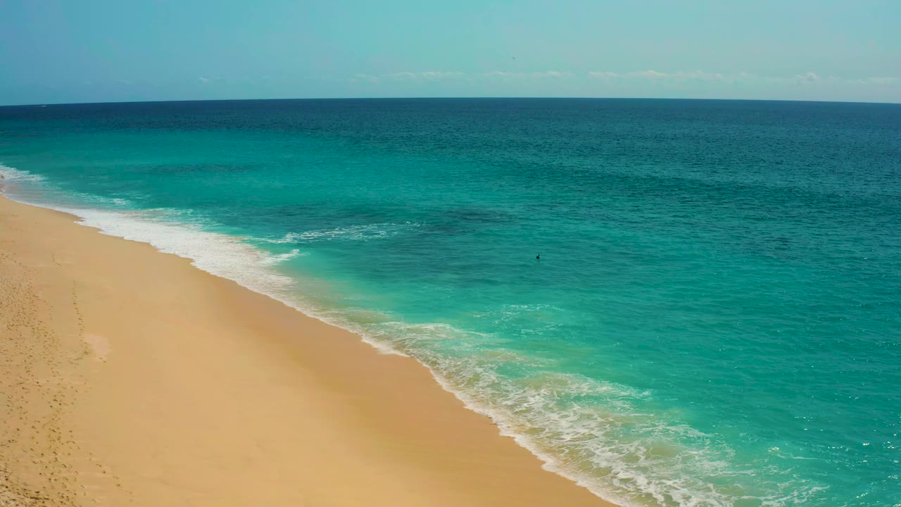 Aerial shoreline frame in Baja California Sur, Mexico, where a curved strip of foam separates dry golden beach from shallow turquoise water, with no visible people.