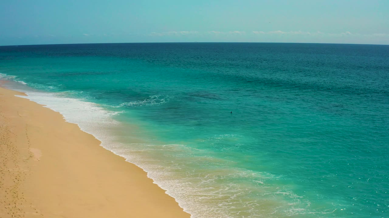 Aerial view over Baja California Sur coastline with a broad band of white foam pushing along the beach edge, clear green-blue water, open sea, and no visible people.