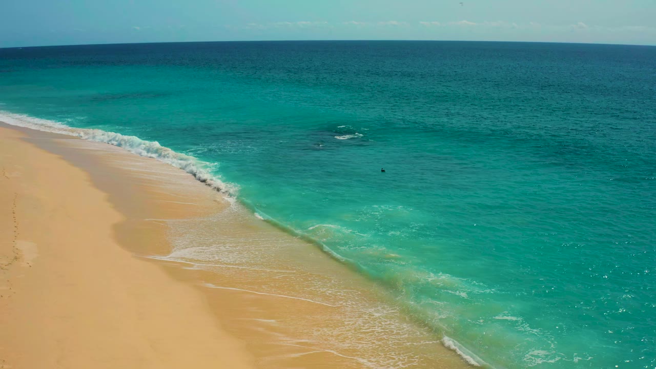 High-angle view over Desert Park Natural Reserve coastline in Baja California Sur, with a thin white wave line crossing the sand and aquamarine water filling the right side, no visible people.