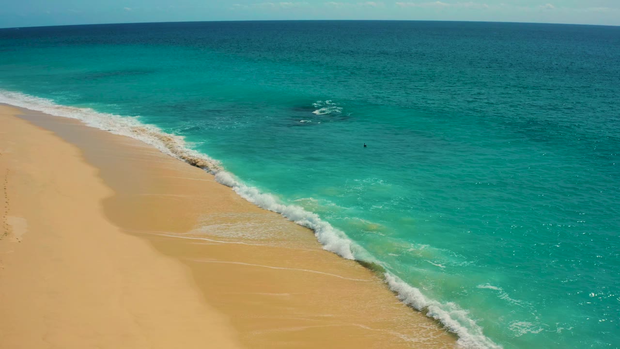Drone-style aerial view in Baja California Sur with a broader white breaker near shore, pale sand on the left, layered turquoise water on the right, and no visible people or structures.