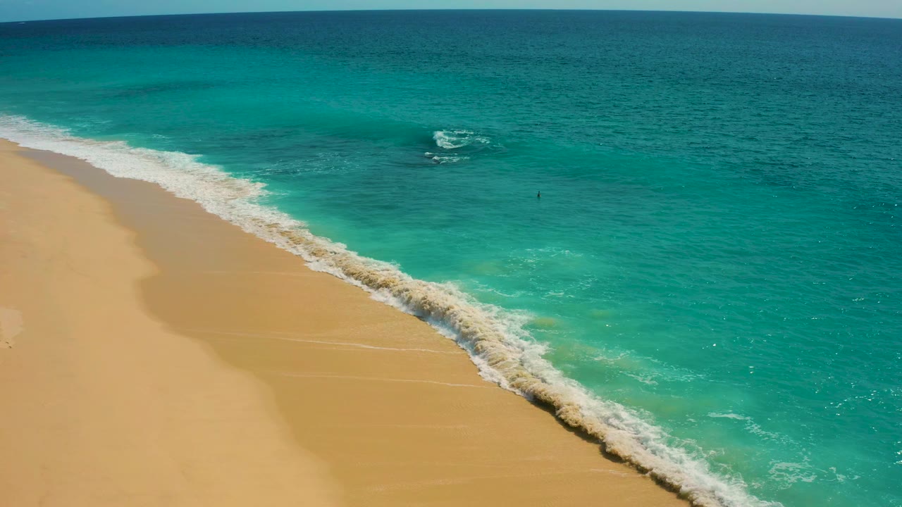 Aerial coastline frame from Desert Park Natural Reserve showing surf sweeping across wet golden sand, clear green-blue shallows, darker sea beyond, and no visible people.