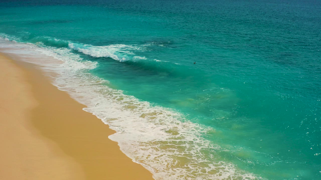 Aerial frame from Desert Park Natural Reserve in Baja California Sur showing turquoise water, white foam, and tan beach with no clear visible person in this opening moment.