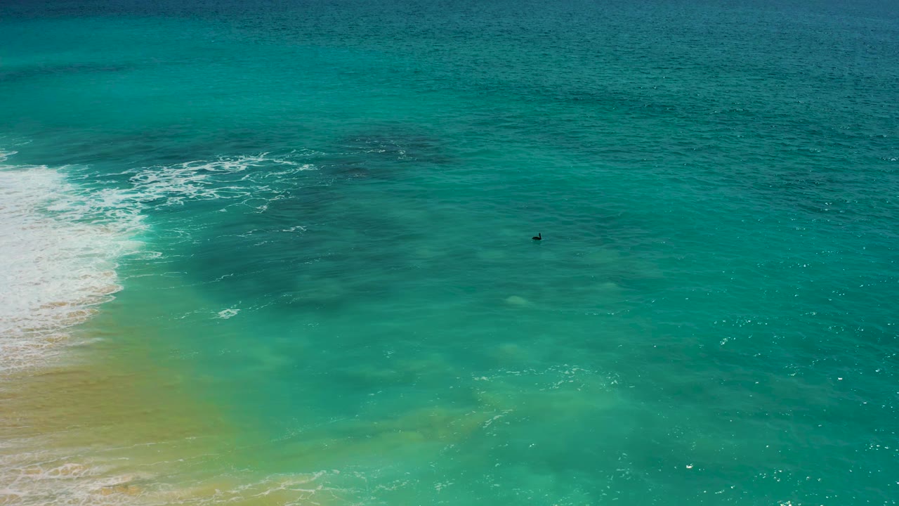 Aerial frame from Baja California Sur showing a band of white foam on the left, translucent green shallows below, and darker teal water above, no visible people.