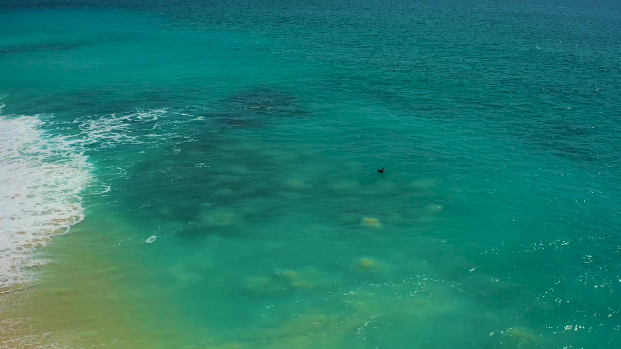 Baja California Sur aerial frame with foamy surf breaking along the sandy left edge and shallow green water spreading toward the center, no visible people.
