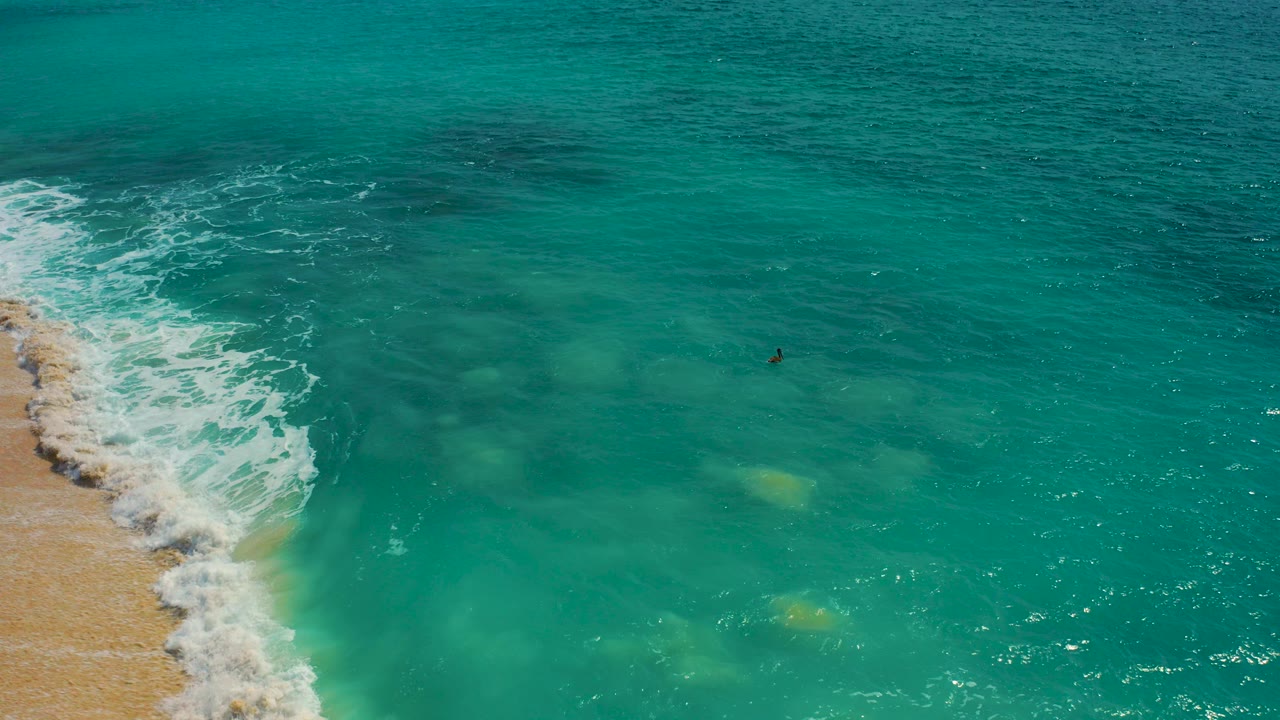 High-angle aerial frame from Baja California Sur shows turquoise water, white waves breaking along the sandy left edge, submerged patches below, and one distant surfer near center.