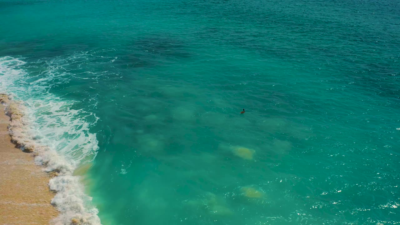 Drone view of a surfer in clear teal water near breaking beach waves.