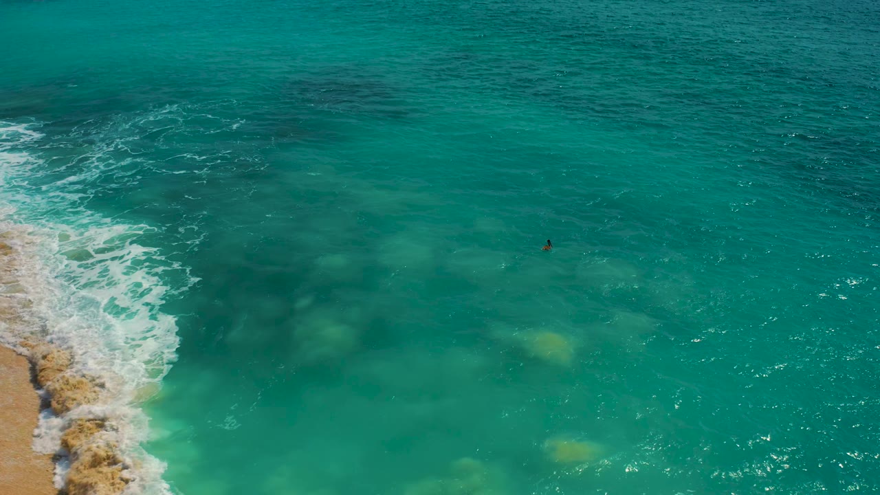 The Baja California Sur aerial view shows the shoreline foam pushed higher along the left edge, with a lone surfer farther out in turquoise water and yellow-green seabed patches below.
