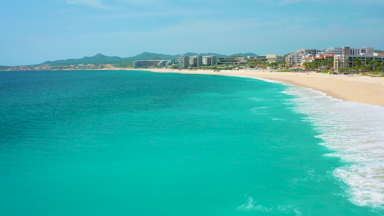 Opening aerial frame over turquoise water and a sandy beach in Desert Park Natural Reserve, Baja California Sur, with modern coastal buildings, low hills, clear sky, and no visible people.