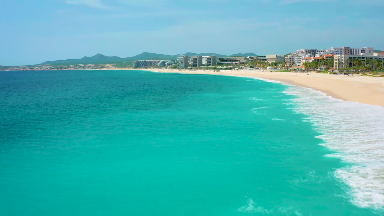 Aerial view one second in showing more open turquoise water before the surf line reaches the sandy shore at Desert Park Natural Reserve, with hotel buildings and hills in the background and no visible people.