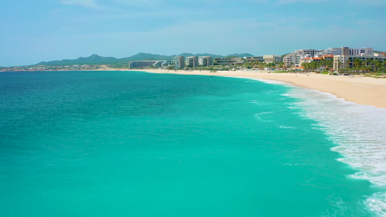 Aerial frame with the turquoise ocean occupying most of the view, a curving white surf band on the right, sandy beach beyond it, resort-style buildings, distant hills, and no visible people.