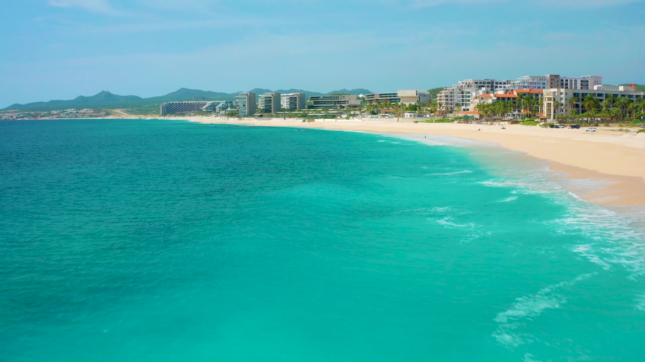 Aerial view of turquoise ocean beside a sandy beach and waterfront buildings.