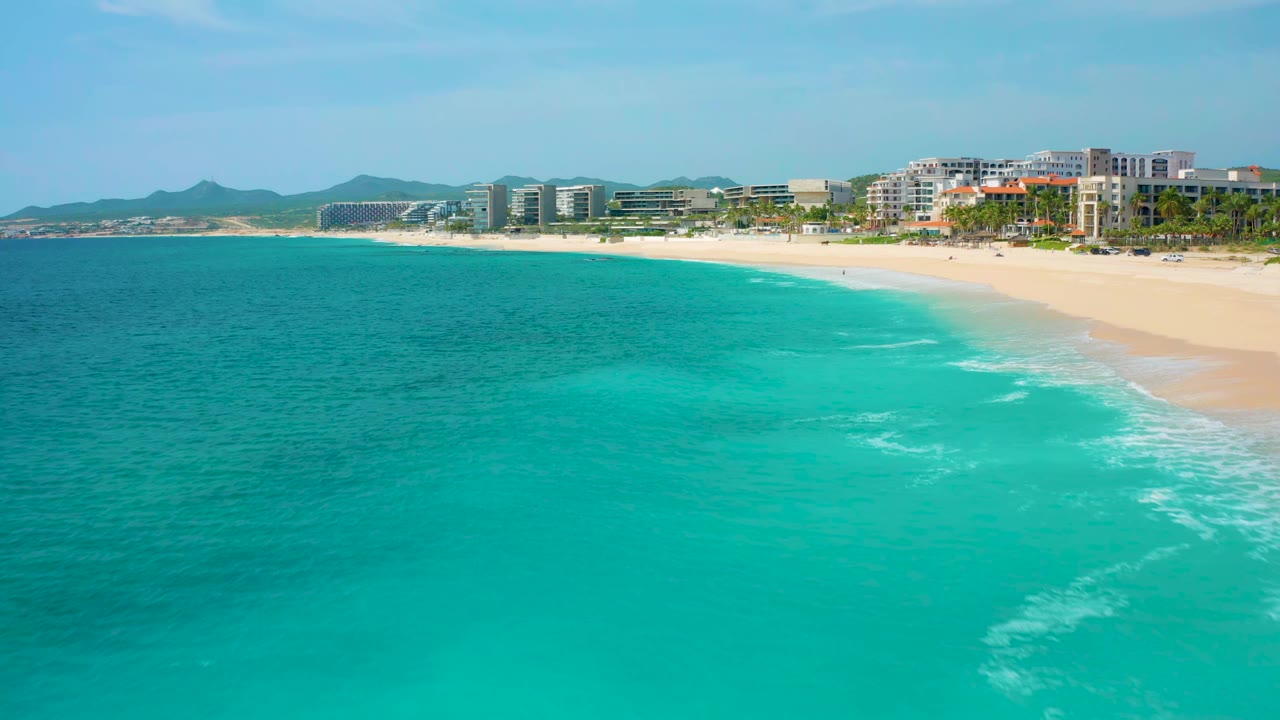 Aerial view one second into the clip over Desert Park Natural Reserve in Baja California Sur, Mexico, showing more open turquoise water, a pale sand shoreline, low white surf, modern coastal buildings, and no visible people.