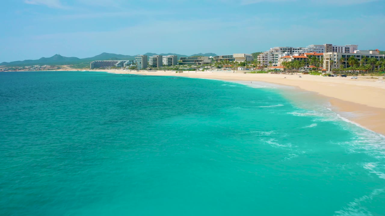 Two seconds in, the aerial frame over Desert Park Natural Reserve in Baja California Sur, Mexico, shows the beach running diagonally along the right edge, turquoise ocean across the foreground, resort-scale buildings, distant hills, and no visible people.