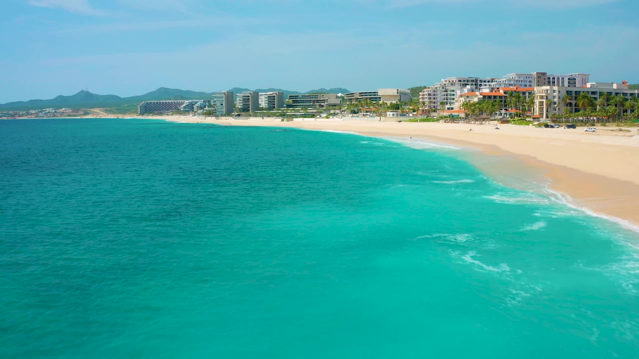 At three seconds, the Desert Park Natural Reserve coastline in Baja California Sur, Mexico, remains in a high aerial view with a broad band of turquoise water, thin surf on the sand, beachfront buildings, hazy hills, and no visible people.