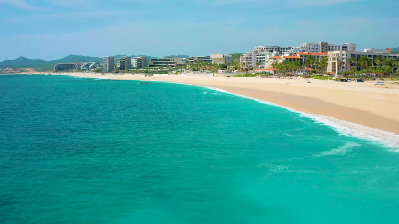 Aerial frame over the Baja California Sur shoreline with shallow turquoise water spreading across the foreground, a curved white wave line near the beach, resort-style buildings in the background, and no visible people.
