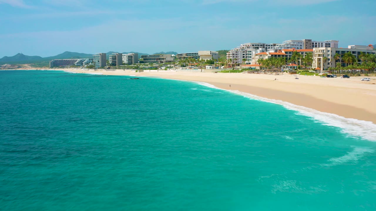Wide aerial view in Baja California Sur, Mexico, showing a larger expanse of turquoise ocean in front of a sandy beach, a line of hotels or residences near the shore, distant hills, and no visible people.