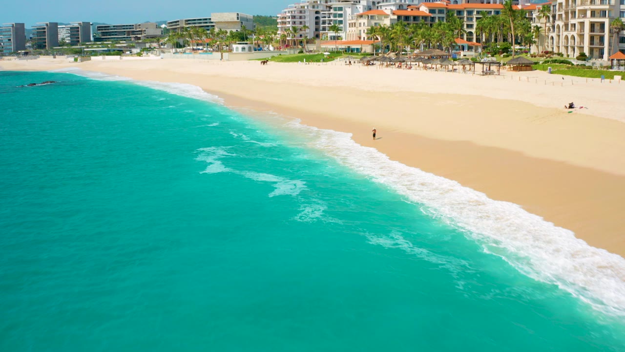 Aerial frame from Desert Park Natural Reserve in Baja California Sur showing turquoise ocean, a broad sandy beach, distant coastal buildings, palms, and a lone person near the shoreline.