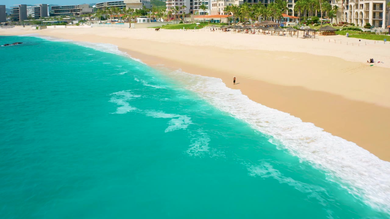 Drone frame from Desert Park Natural Reserve showing a diagonal line of surf, open pale sand, turquoise water, and a single distant walker close to the breaking waves.