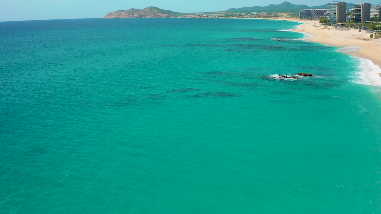 High-angle aerial frame over Desert Park Natural Reserve in Baja California Sur, showing turquoise water, a sandy beach at upper right, distant buildings, mountains, and no visible people.