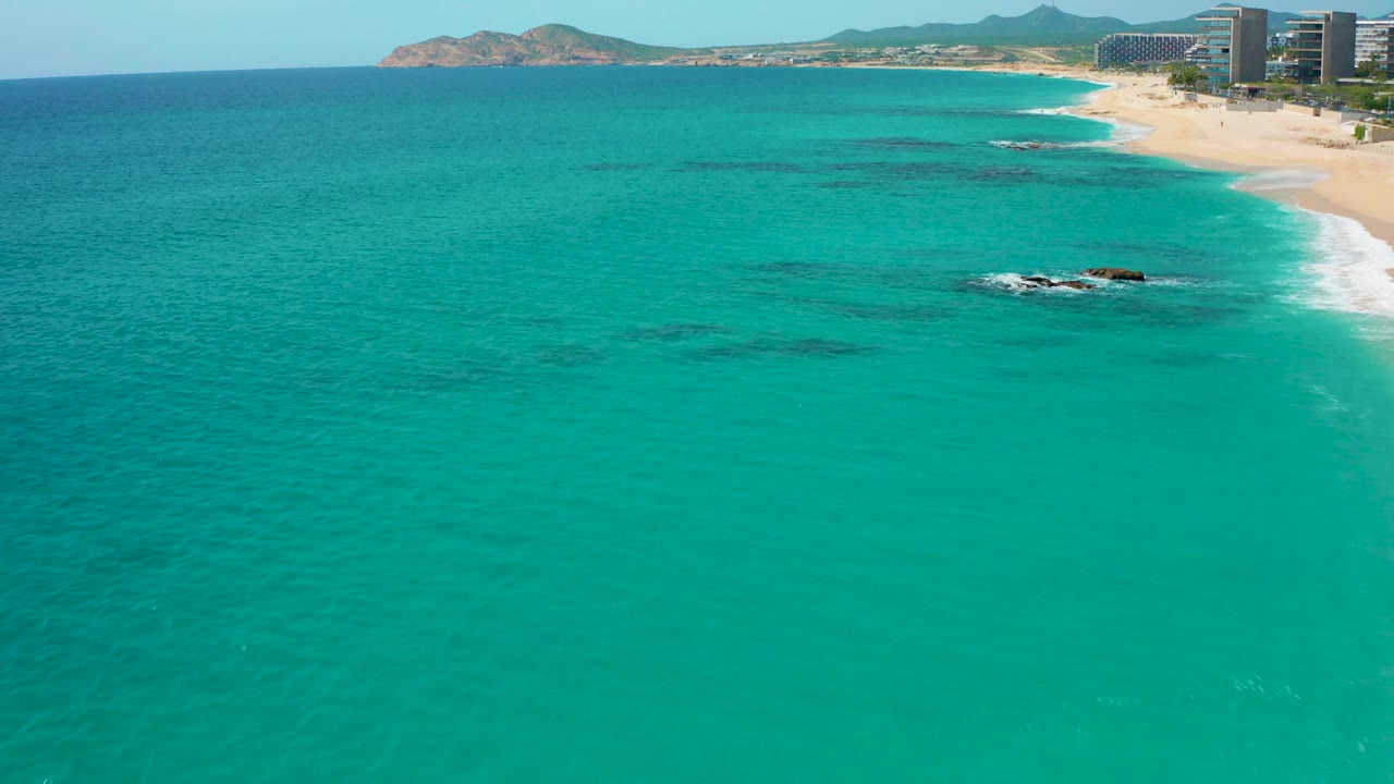 Aerial frame in Baja California Sur with clear turquoise ocean across the foreground, dark underwater formations near center, a sandy shoreline at upper right, distant buildings, and no visible people.
