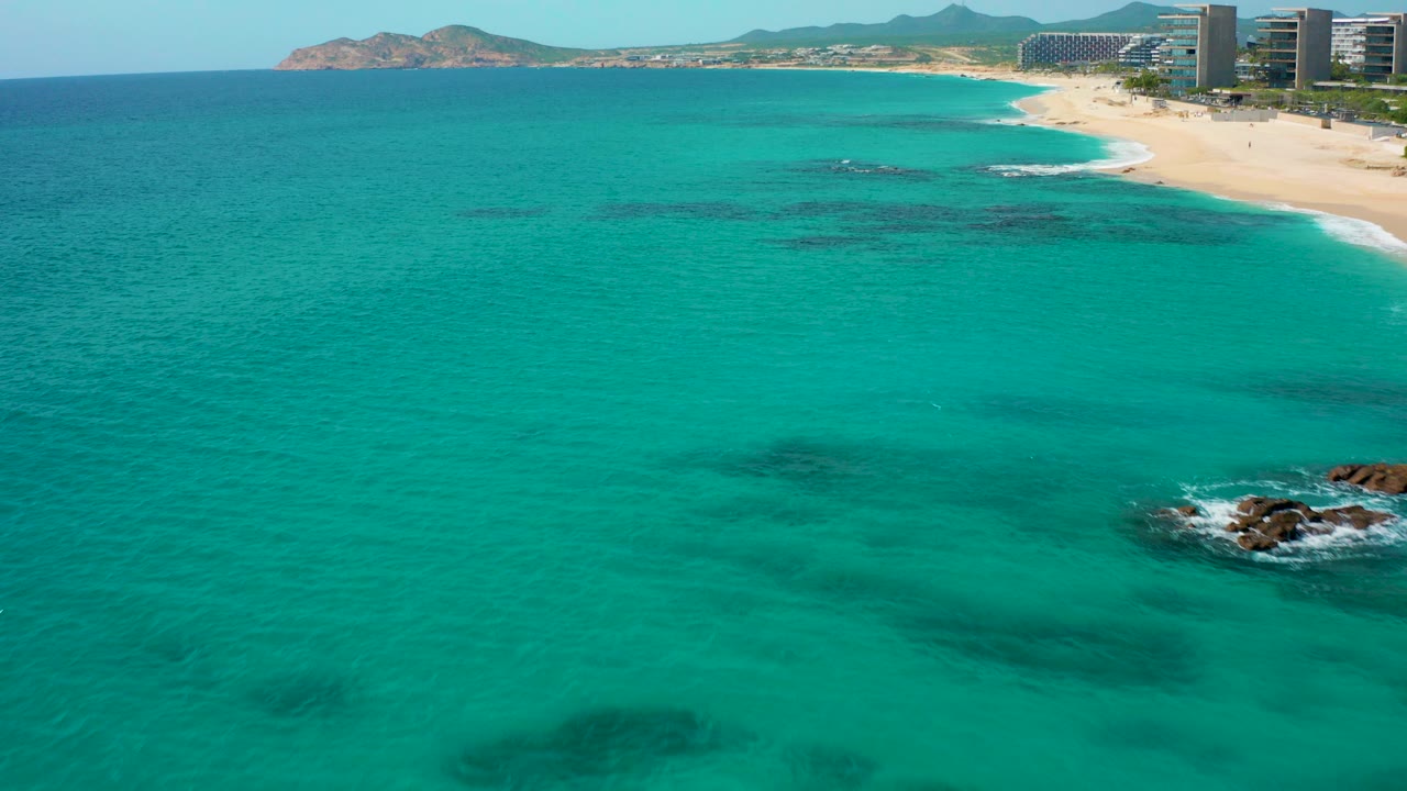 High aerial view over turquoise water in Desert Park Natural Reserve, Baja California Sur, with a sandy beach, coastal towers, distant hills, and no visible people.