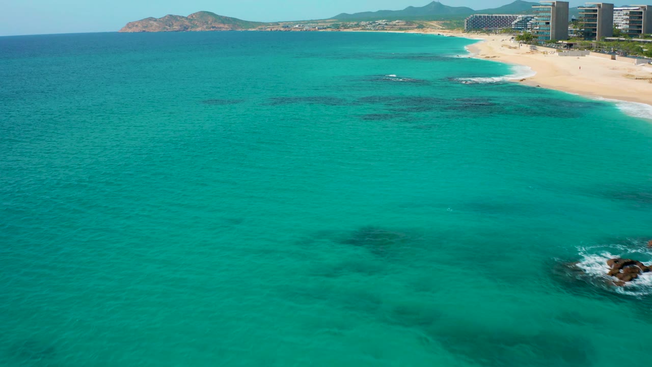 High drone view across turquoise water in Baja California Sur, with darker underwater areas, a narrow sandy shoreline, distant high-rises, low hills, and no visible people.