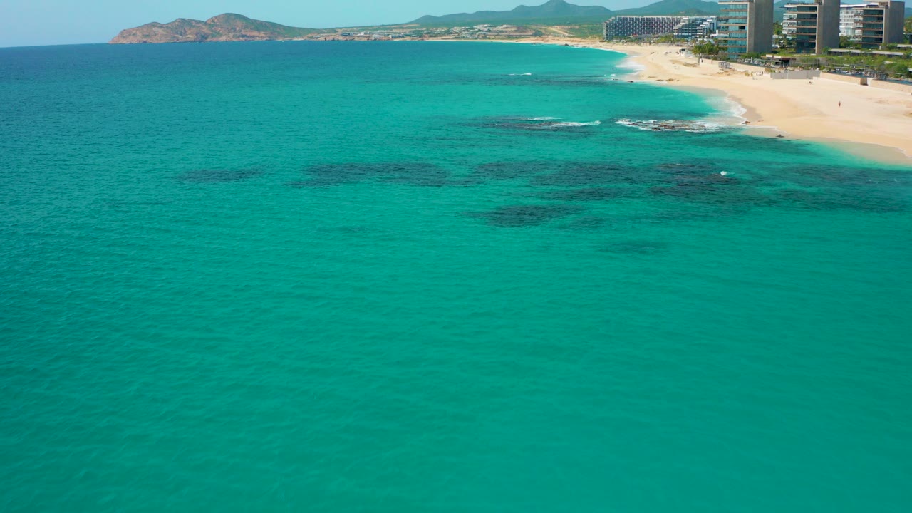 Opening aerial frame over Desert Park Natural Reserve in Baja California Sur, Mexico, with turquoise water, dark submerged patches, a sandy shoreline, distant hills, beachfront buildings, and no visible people.