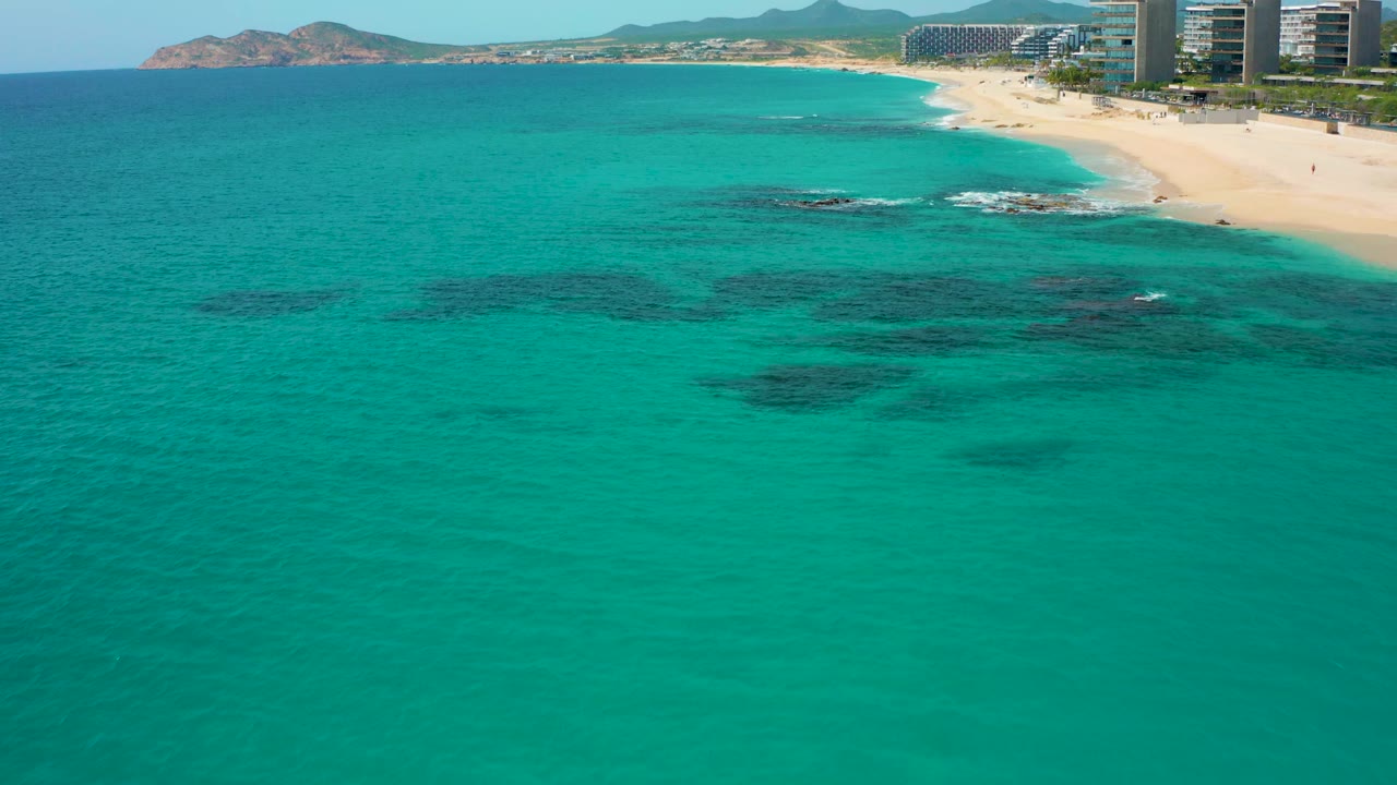 At 0:03, the drone view shows clear blue-green ocean in front, darker underwater formations, a sandy beach with white wash, modern buildings along the shore, hills behind, and no visible people.
