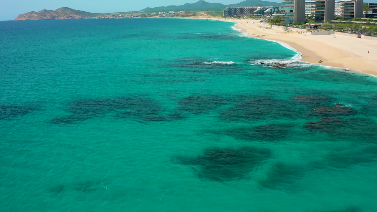 Opening frame of Desert Park Natural Reserve in Baja California Sur, Mexico, showing turquoise water, dark underwater patches, pale sand, distant beachfront towers, hills, and no visible people.