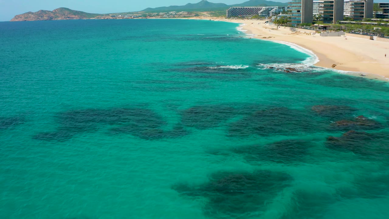 At 0:02, the frame shows a broad turquoise foreground, a thin line of surf on the sand, modern high-rises along the shore, distant hills, and no visible people.