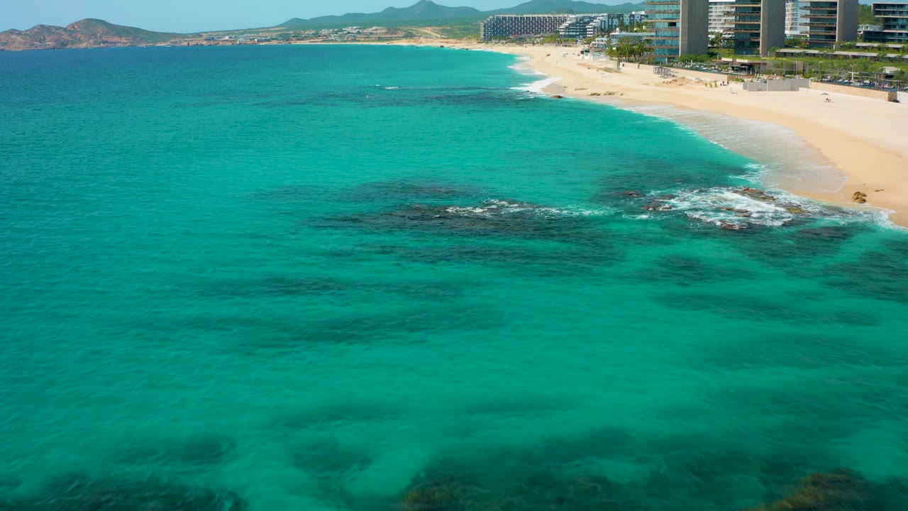 High aerial view over turquoise water at Desert Park Natural Reserve in Baja California Sur, with dark submerged rocks, sandy beach, distant hills, hotel towers, and no visible people.
