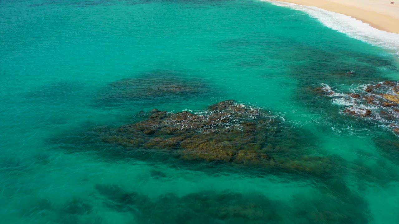 High-angle aerial view in Baja California Sur showing turquoise water over dark rock formations, a pale sandy beach along the upper edge, white foam on the rocks, and no visible people.