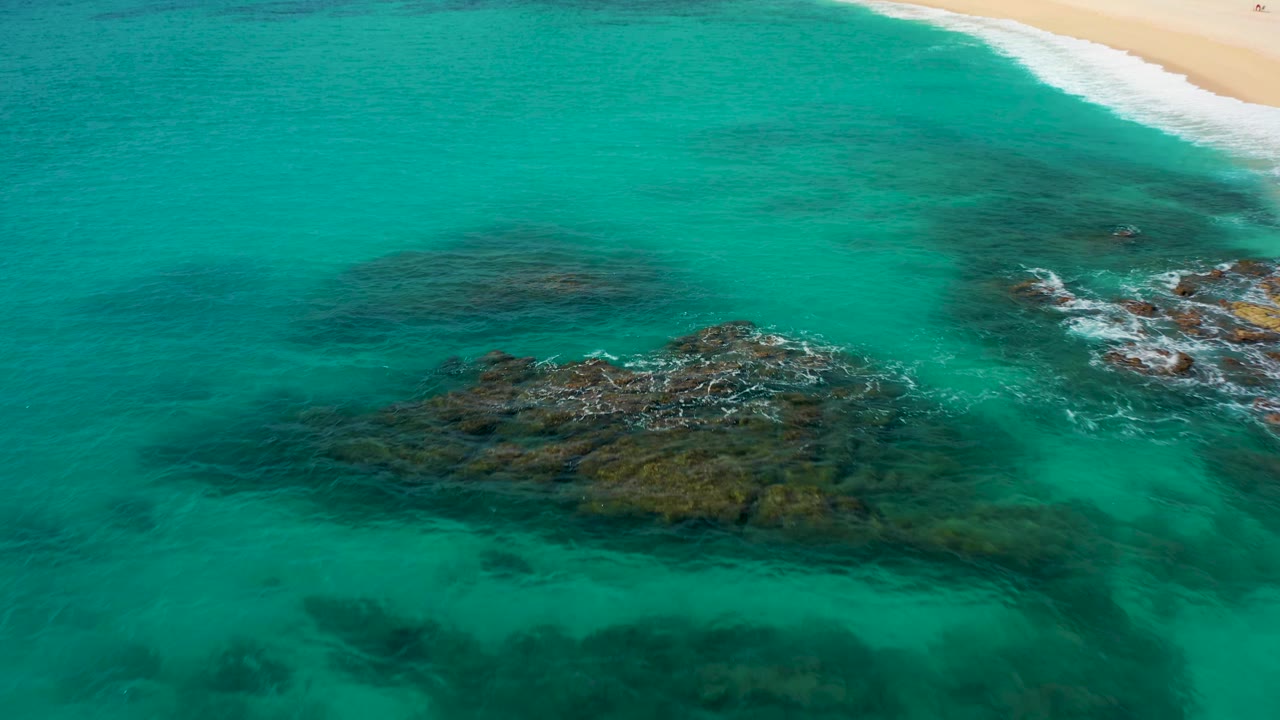 Aerial frame over Baja California Sur shallows with a central rock cluster, transparent turquoise water, white surf streaks, a narrow beach strip at the top, and no visible people.