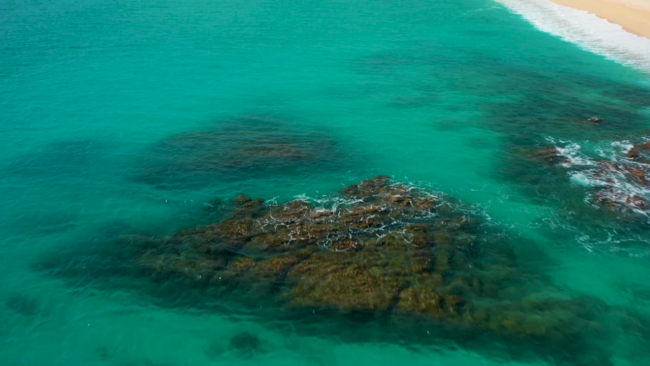 High-angle drone view in Baja California Sur with a larger dark rock mass in turquoise water, small white wave breaks on the right, beach surf near the upper edge, and no visible people.