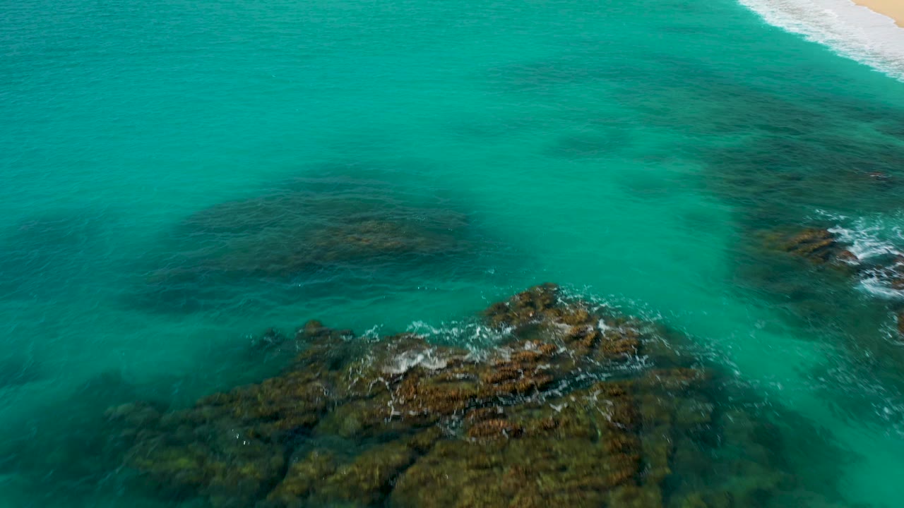 Aerial view over Baja California Sur water showing submerged rock patches across turquoise shallows, exposed rocks with foam in the lower right, a small beach edge at the top right, and no visible people.
