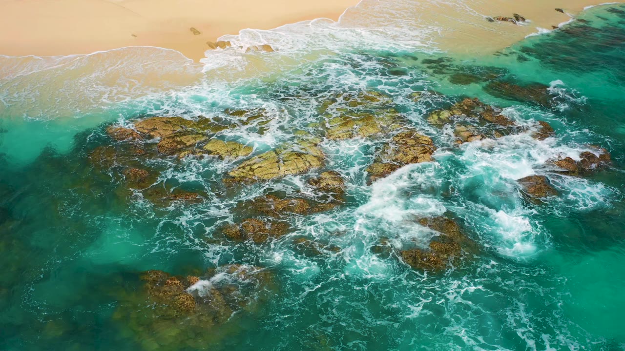 Overhead view in Desert Park Natural Reserve, Baja California Sur, Mexico, with surf curling around clustered rocks and the sandy beach running along the upper frame, no visible people.