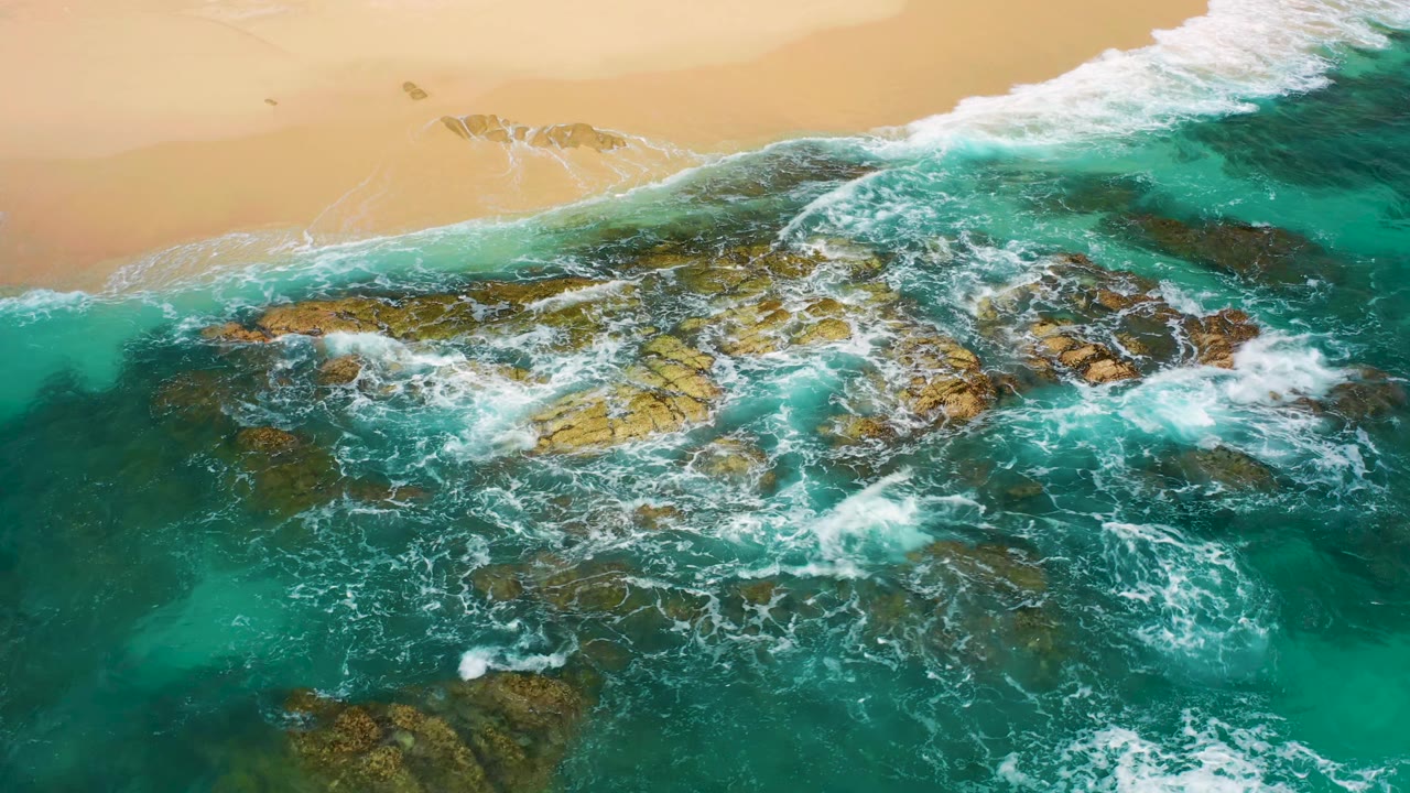 Aerial shoreline frame from Desert Park Natural Reserve with white water spreading across shallow brown-green rock shelves and a pale sand band at the top, no visible people.