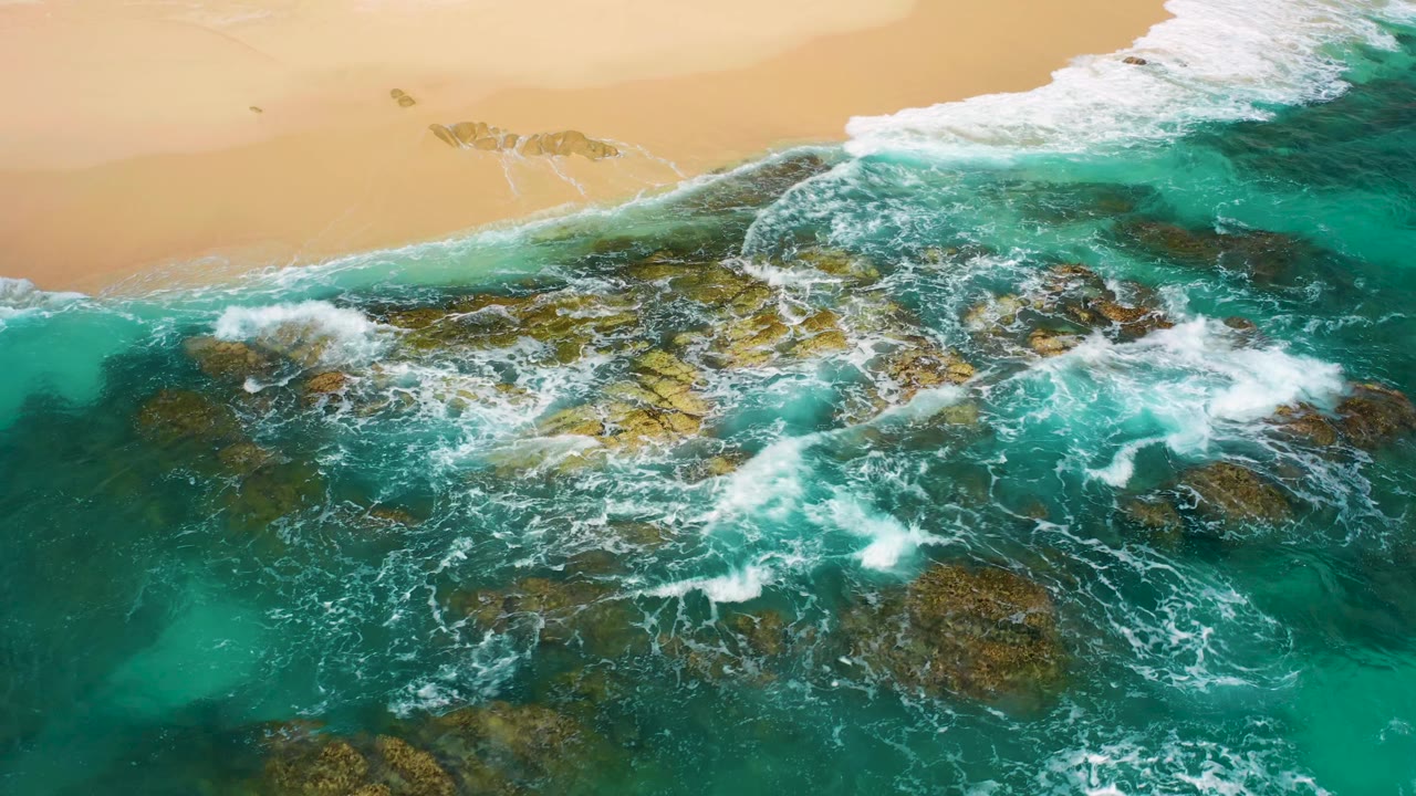 Top-down Baja California Sur coast detail with a larger band of white wash crossing jagged rocks in clear blue-green water, with sand visible along the upper edge and no visible people.