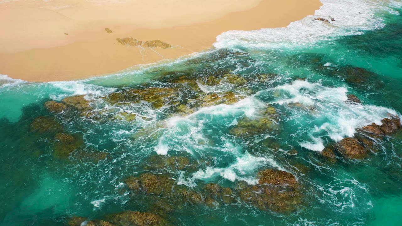 Aerial drone-looking frame over Desert Park Natural Reserve shoreline where foamy turquoise surf covers a broad rocky ledge below a warm sand beach, no visible people.