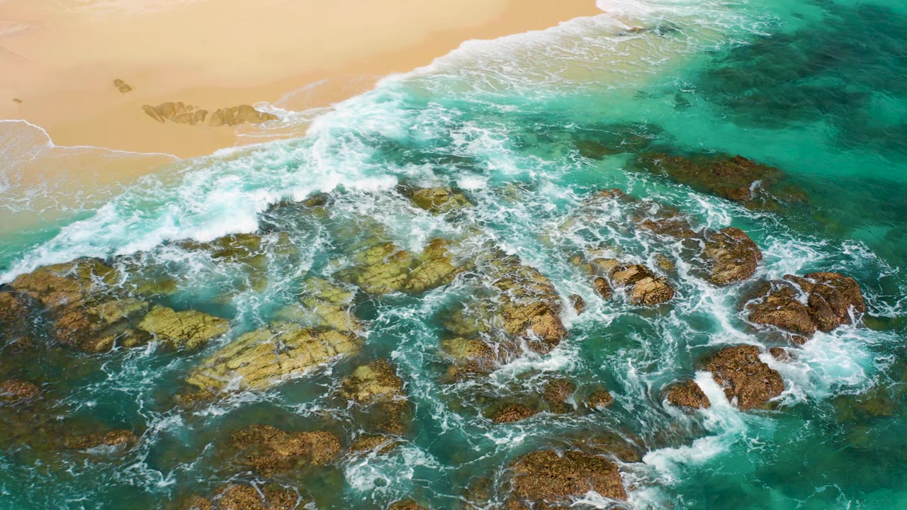 Top-down aerial view of turquoise waves breaking over jagged rocks beside golden sand in Desert Park Natural Reserve, Baja California Sur, with no visible people.
