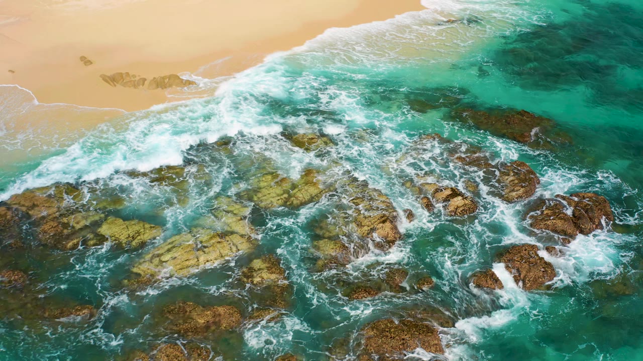 Aerial frame of white foam spreading through clear turquoise water around tan and brown rock formations in Desert Park Natural Reserve, Baja California Sur, with no visible people.