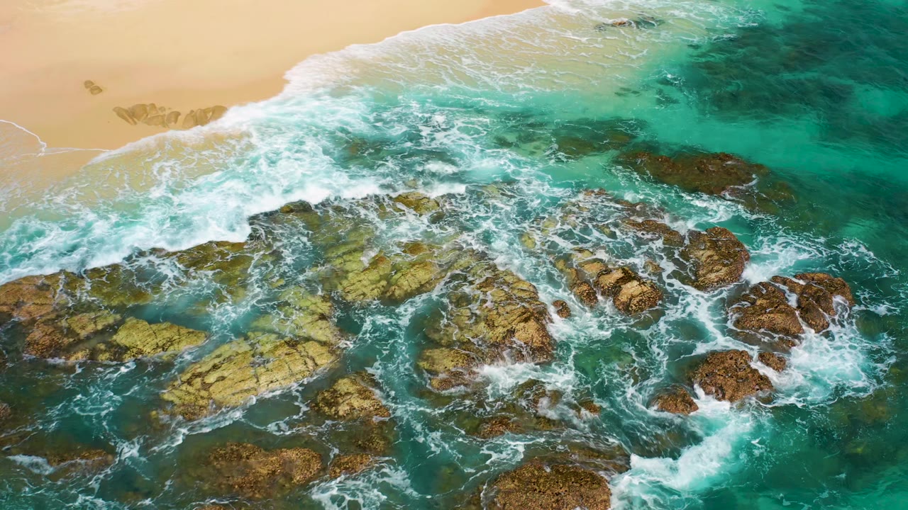 Top-down drone view of waves crossing a rocky reef below a smooth sandy beach edge in Desert Park Natural Reserve, Baja California Sur, with no visible people.