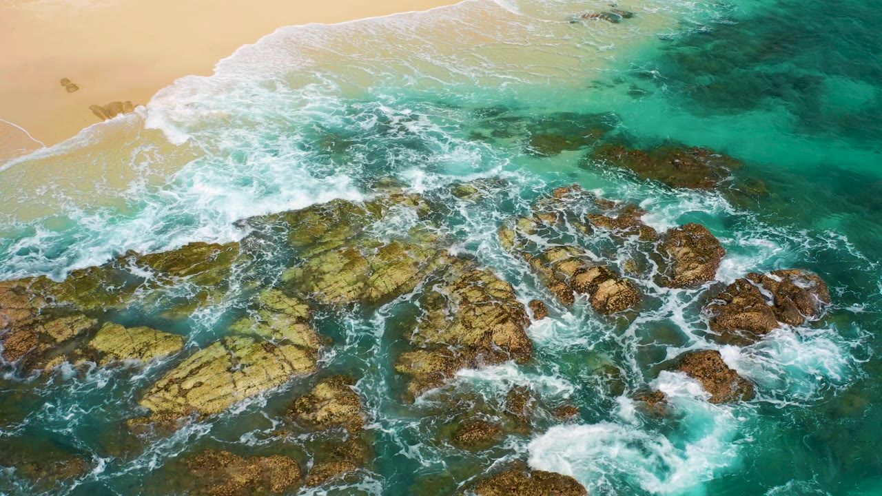Aerial view of a diagonal surf line washing over larger coastal rocks and green-blue water in Desert Park Natural Reserve, Baja California Sur, with no visible people.
